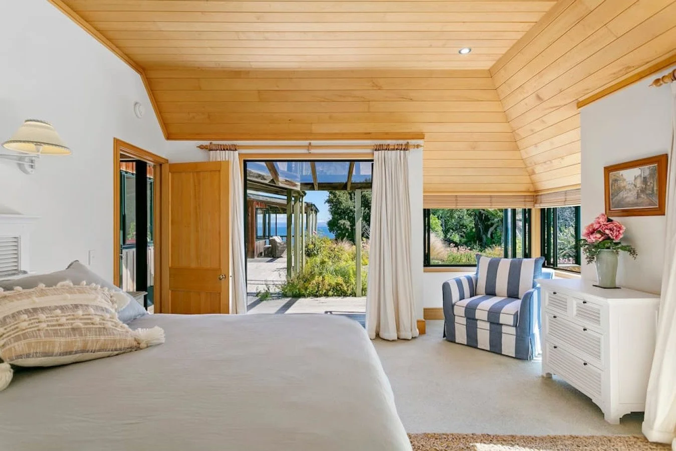 A cozy bedroom with a wooden ceiling, white walls, and a large window showing a garden and outdoor patio. The room has a white bed with pillows, a striped blue and white armchair, and a white dresser with a flower vase. There are curtains and a frame