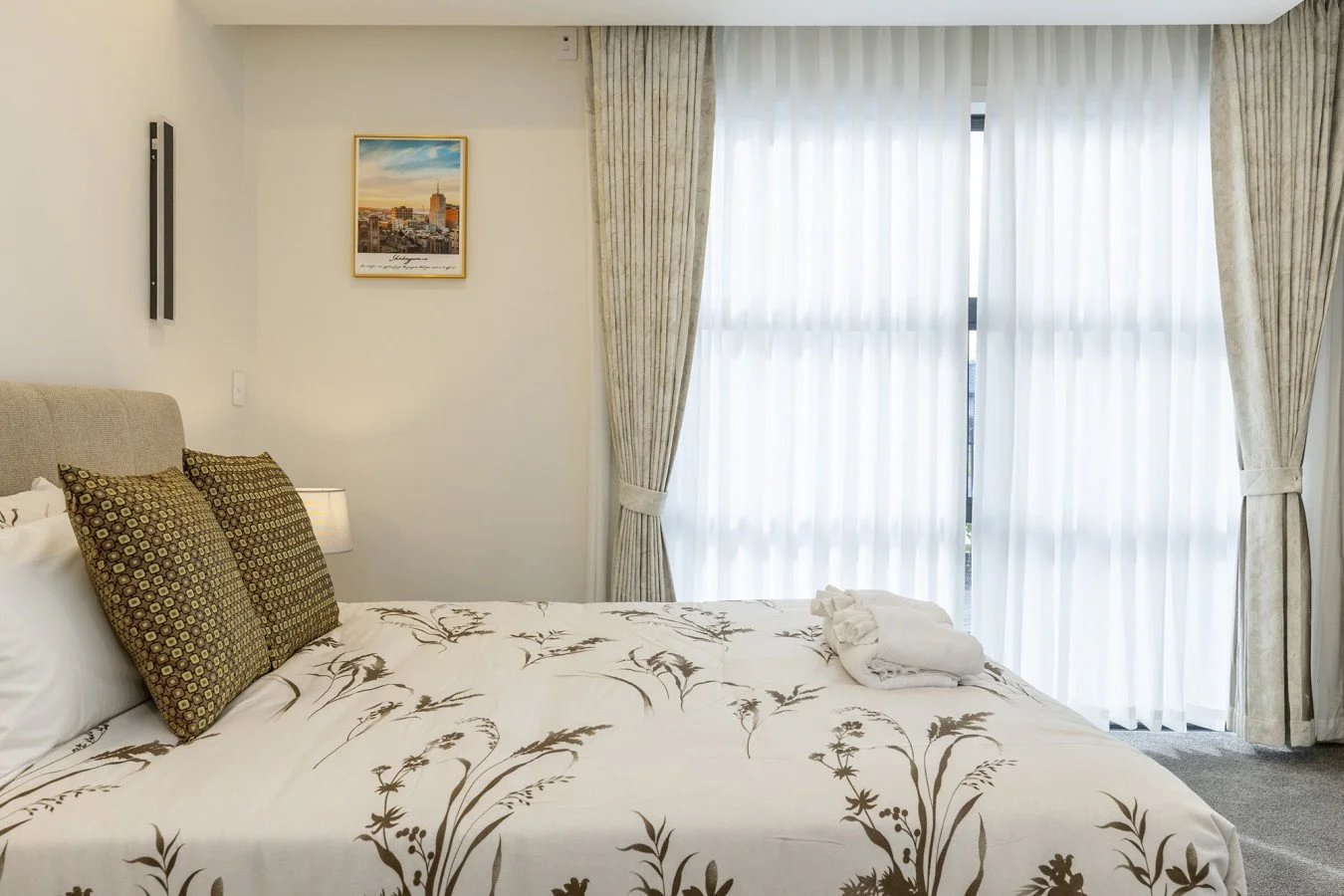 A neatly made hotel bed with two decorative patterned pillows, a bed with floral-printed sheets, a bedside lamp, and a window with sheer curtains and drapes.