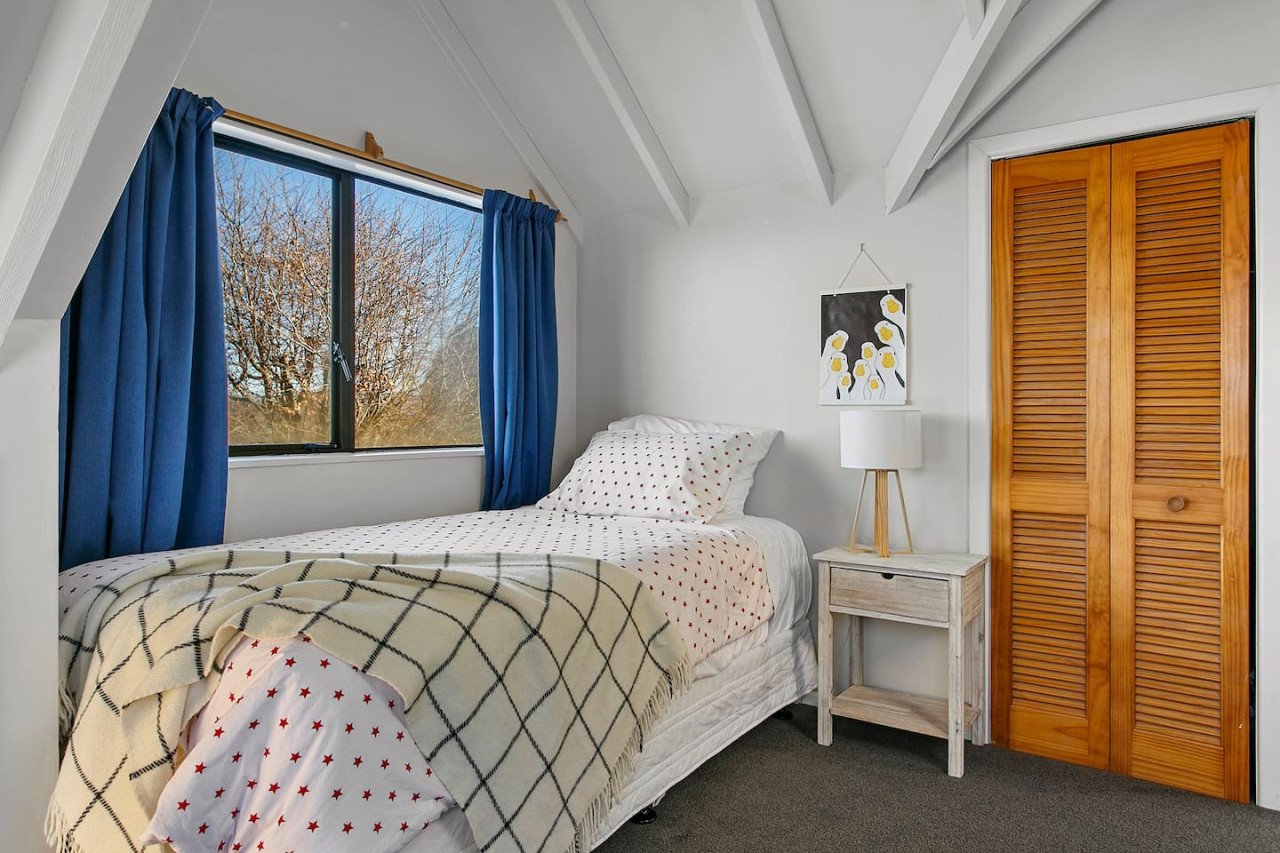 A cozy bedroom with a bed, bedside table, lamp, window with blue curtains, and wooden closet doors.