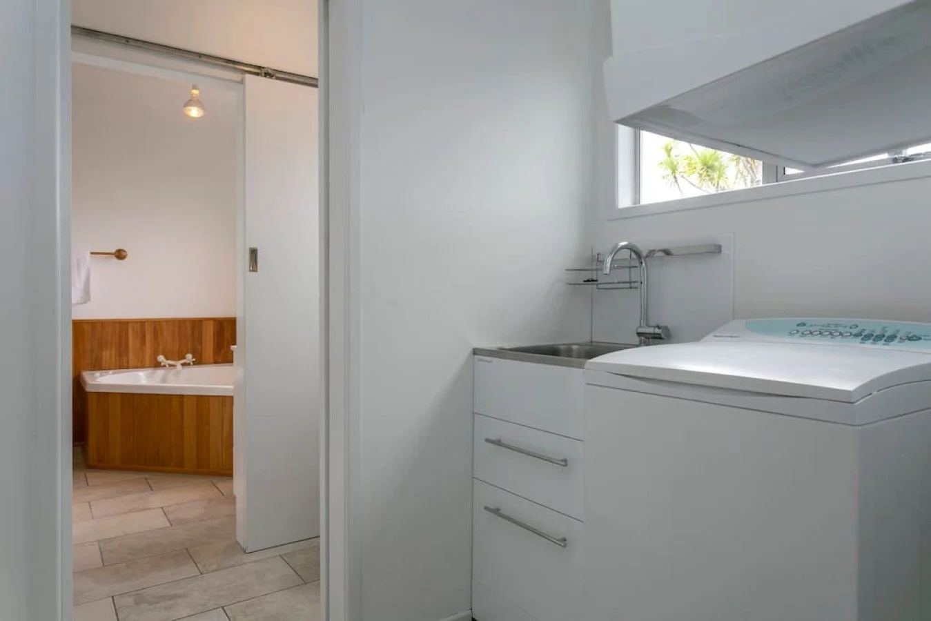 Small laundry area with a washing machine and sink, next to a window, and a bathroom with a bathtub visible through a doorway.