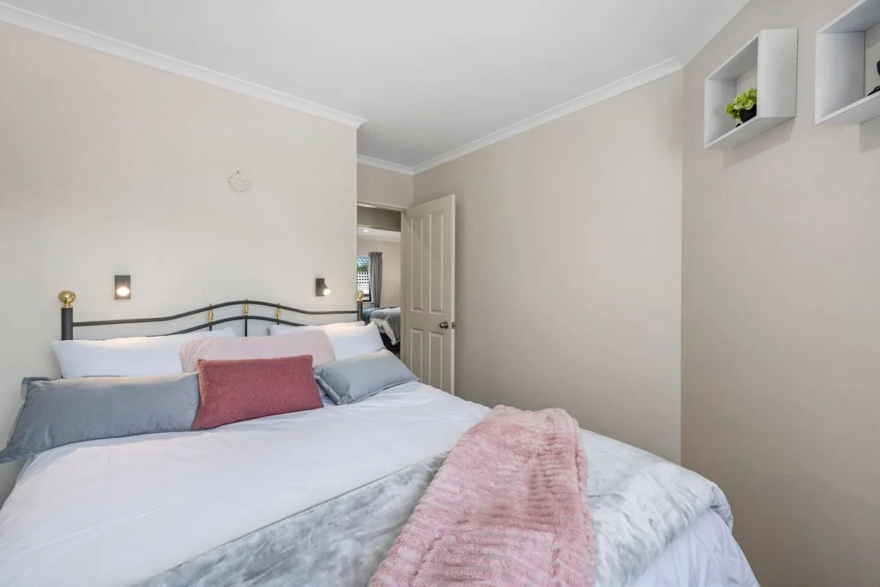 A neatly made bed with multiple pillows and a pink throw blanket in a bedroom, with a door open revealing another room in the background.