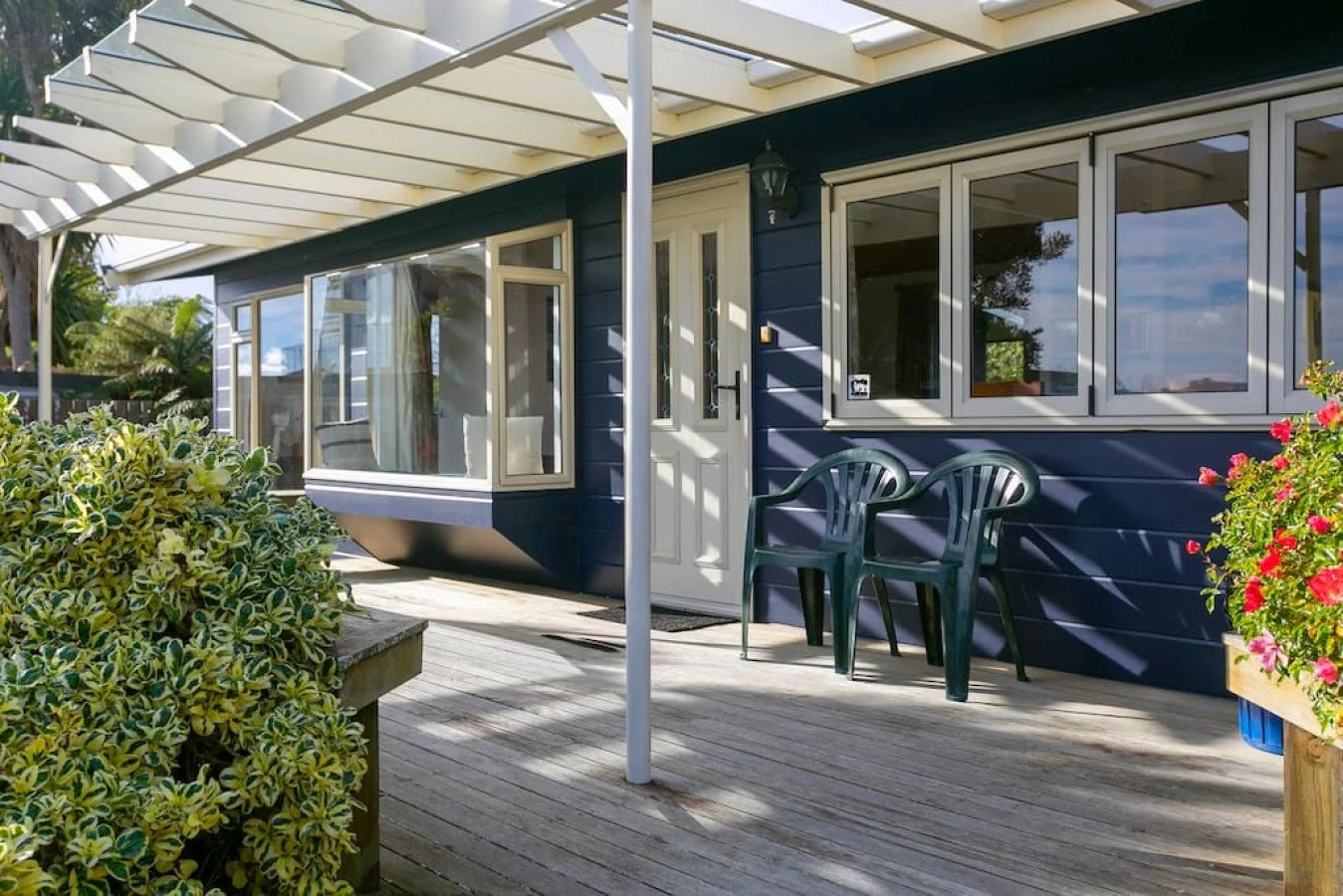 A house with a wooden deck, blue siding, large windows, a white door, and outdoor plastic chairs, with potted plants and bright sunlight.