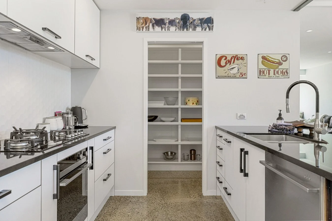 Modern kitchen with white cabinets, black countertops, open shelving, and vintage-style wall signs for coffee and hot dogs.
