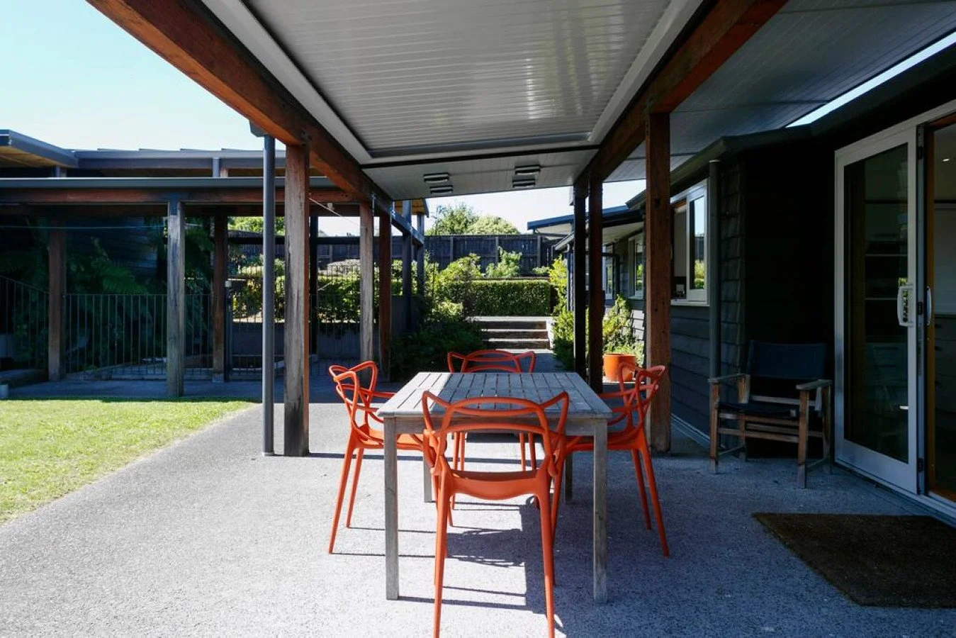 Outdoor patio with a rectangular wooden table and six orange chairs, covered by a roof attached to a house with dark siding and sliding glass door.