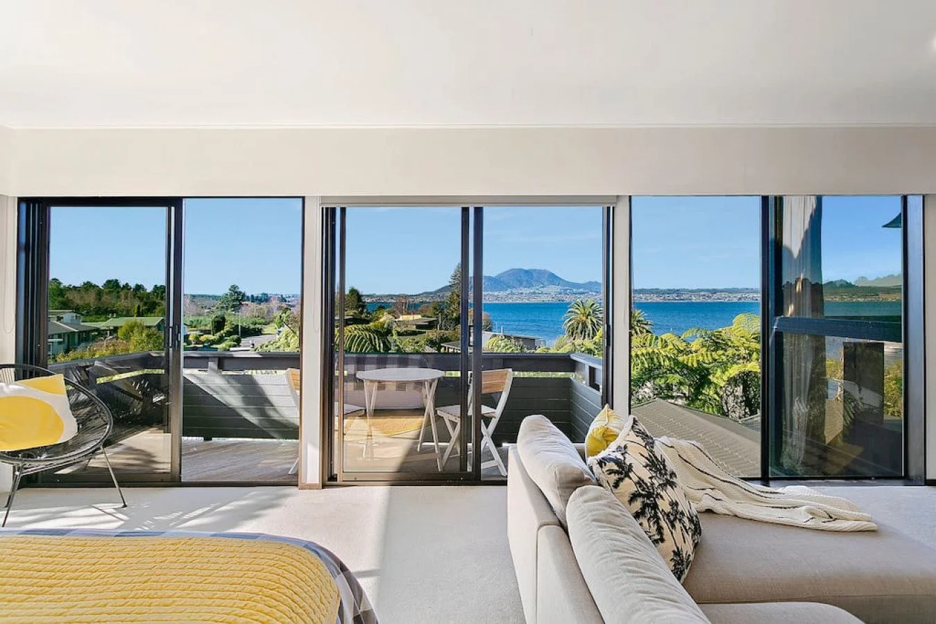 Living room with large glass sliding doors leading to a balcony overlooking a body of water, mountains, and greenery.