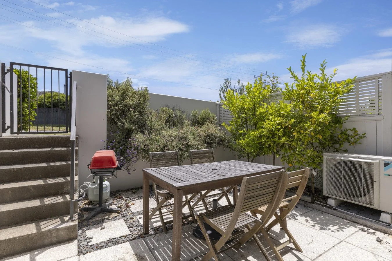 Patio with wooden table and chairs, green shrubbery, air conditioning unit, and gas grill under a blue sky with clouds.