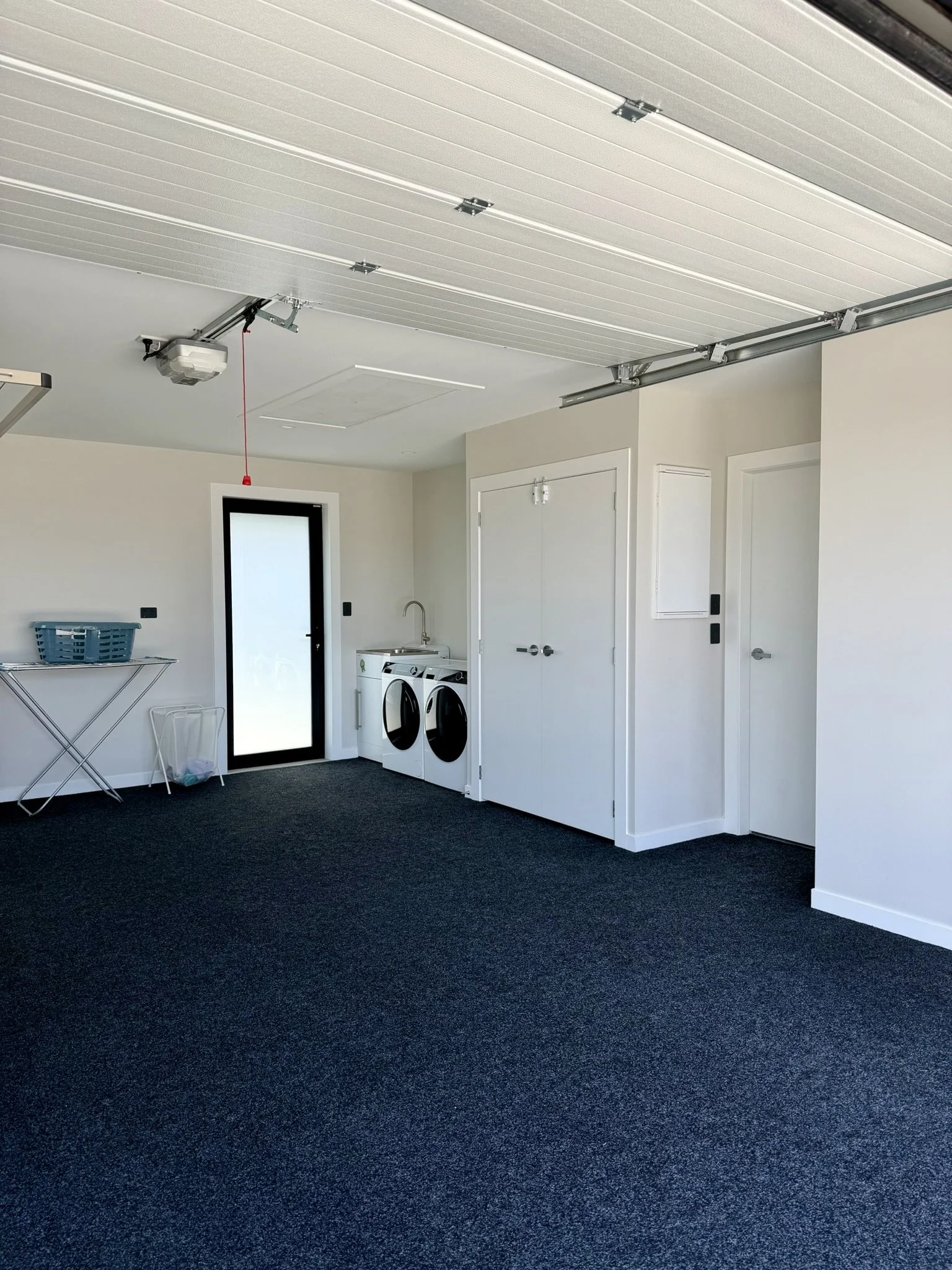 Clean, empty garage with a black carpeted floor, white walls, a laundry area with a washer and dryer, a small sink, a door with frosted glass leading outside, a closed storage closet, and a garage door opening mechanism with a red cord hanging from i