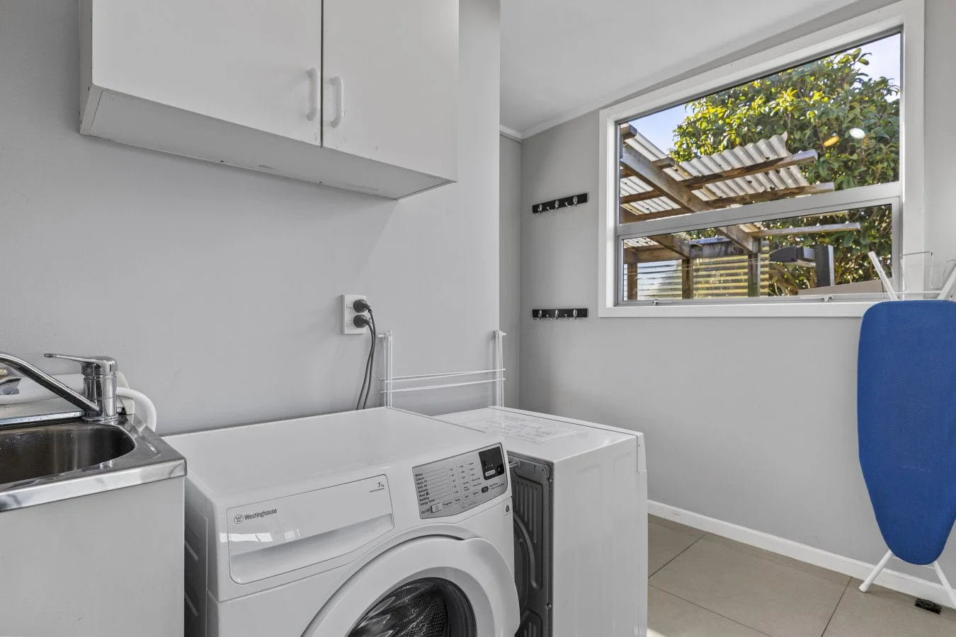 Laundry room with washing machine, small sink, wall-mounted cabinet, and window showing outdoor patio with plants and wooden pergola.