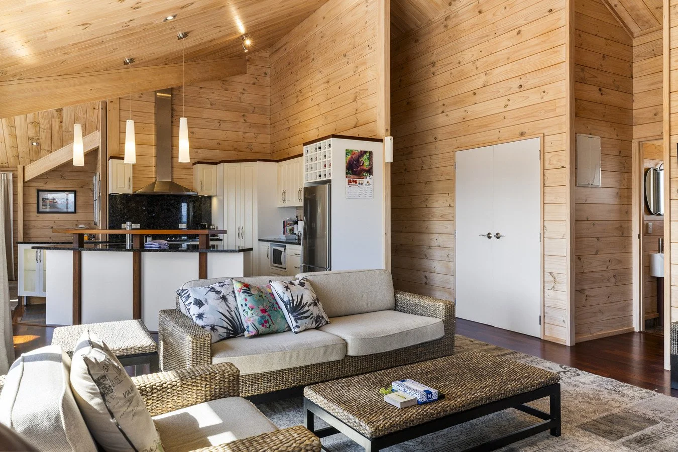 Cozy living room with woven sofa and armchair, wooden walls and ceiling, modern kitchen in background with white cabinets and black countertops, decorative pillows on sofa, and a wooden coffee table.
