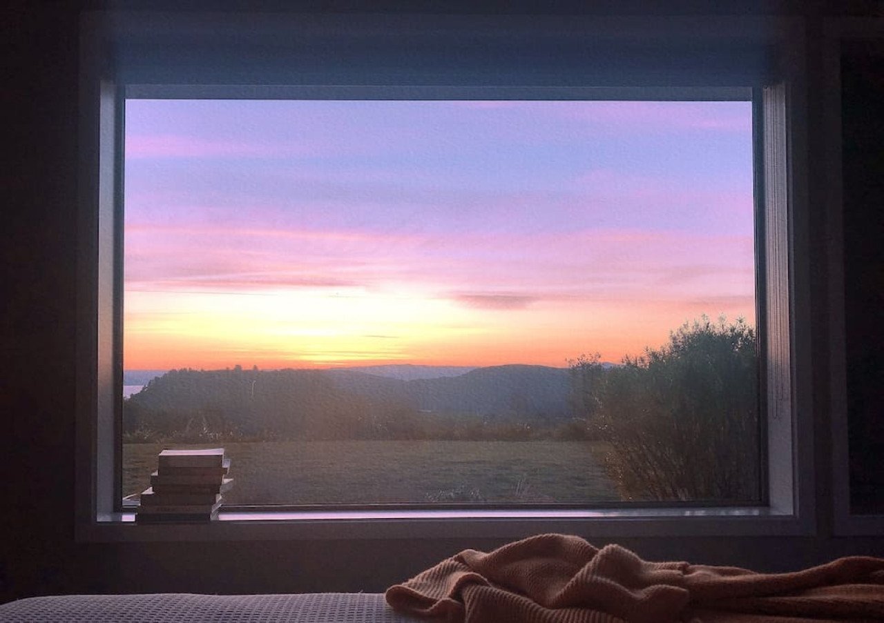Sunset view through a window with a mountain landscape and colorful sky, seen from indoors with a blanket in the foreground.