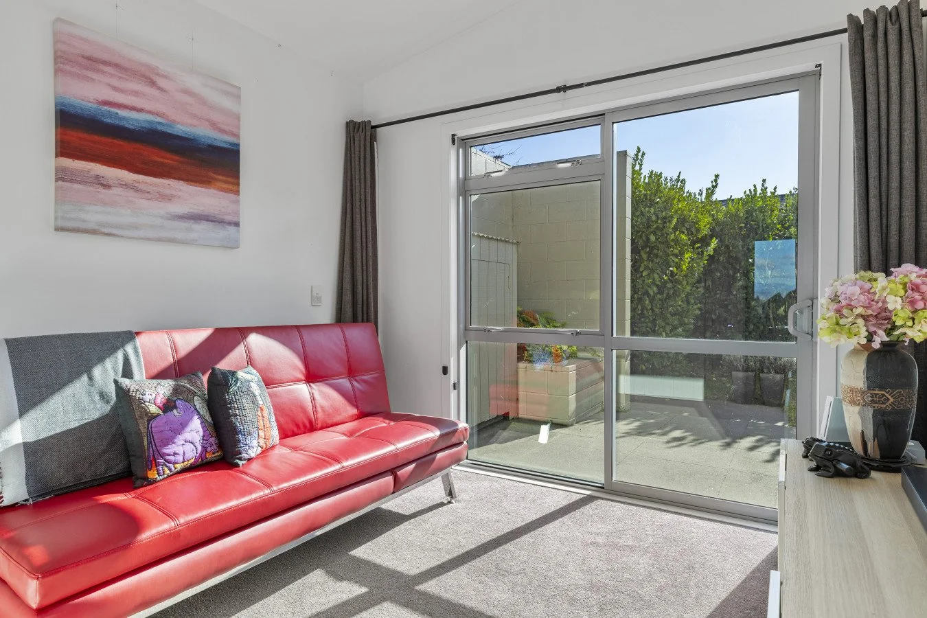 Living room with red leather couch, colorful pillows, wall art, large sliding glass door, and view of patio with potted plants outside.