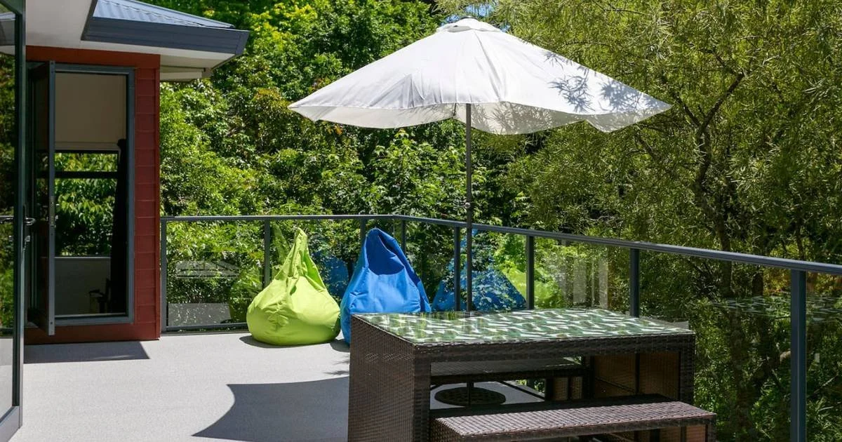 Balcony with a white patio umbrella, two bean bag chairs (green and blue), a dining table with a glass top, and a glass railing with a lush green forest in the background.