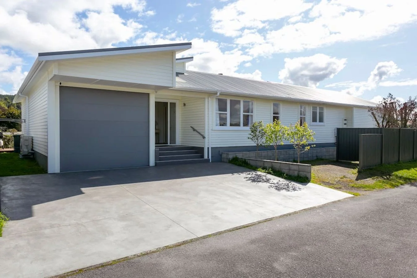 Front view of a white house with a gray garage door and a concrete driveway, blue sky with some clouds, and small front yard with plants and trees.