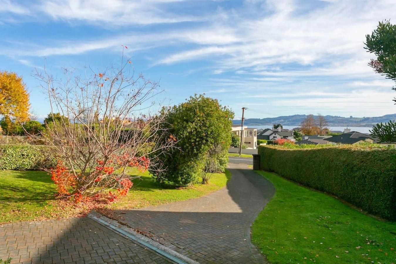 A residential neighborhood with a curved brick-paved driveway, green lawn, trees with fall foliage, a hedge, and houses overlooking a body of water under a blue sky with clouds.