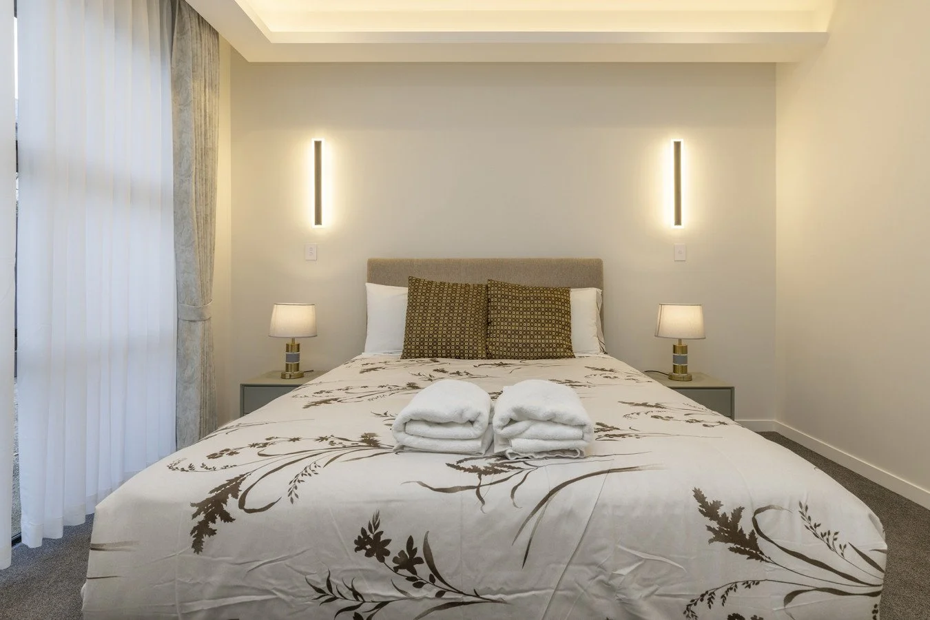 Modern bedroom with a bed, two bedside tables with lamps, two pillows, and folded towels on the bed.