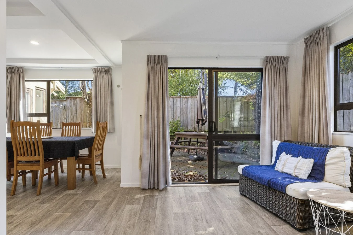 Living room with beige curtains, a wicker sofa with blue cover, a small white side table, wooden dining table with chairs, and a sliding glass door leading to a patio with outdoor furniture and a wooden fence