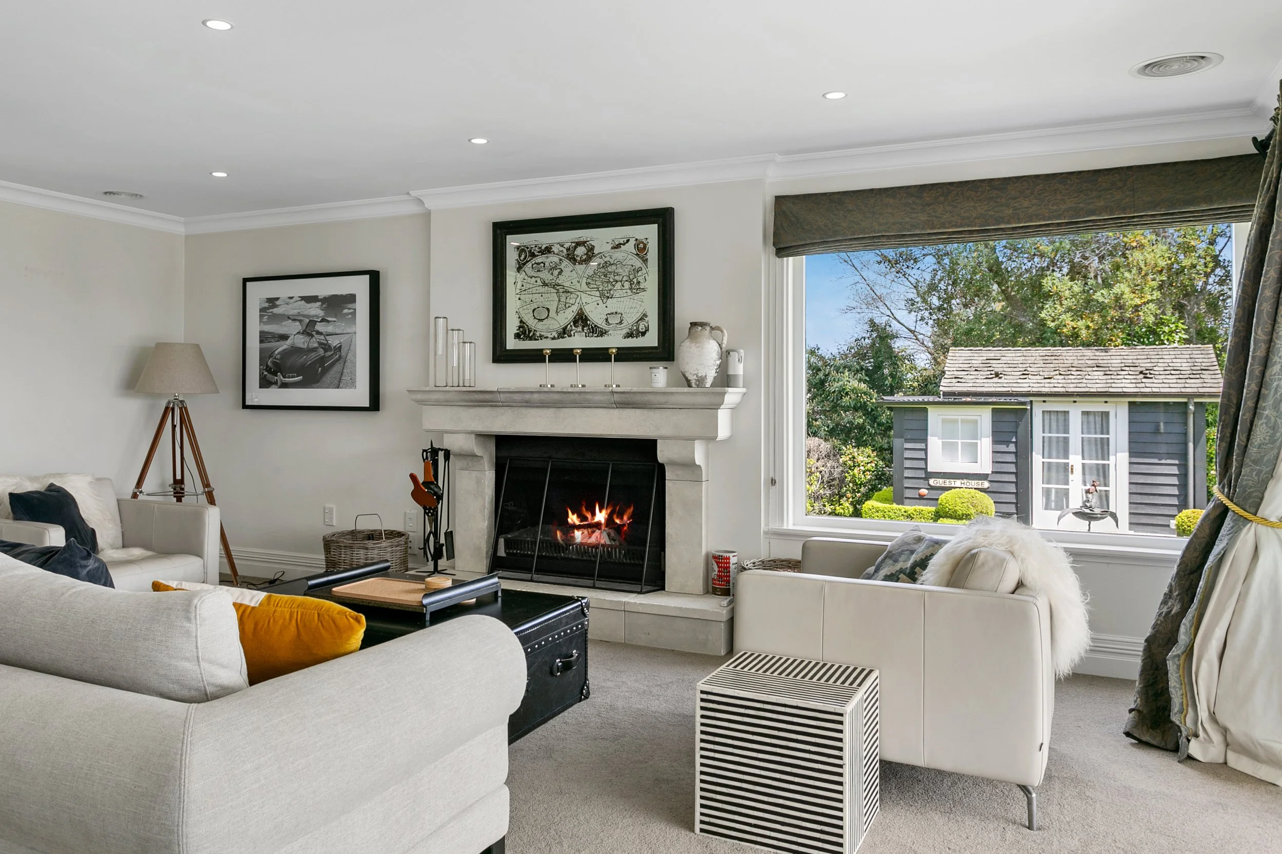 Living room with white walls, fireplace, large window showing a guest house outside, and furniture including sofas, a coffee table, and a lamp.