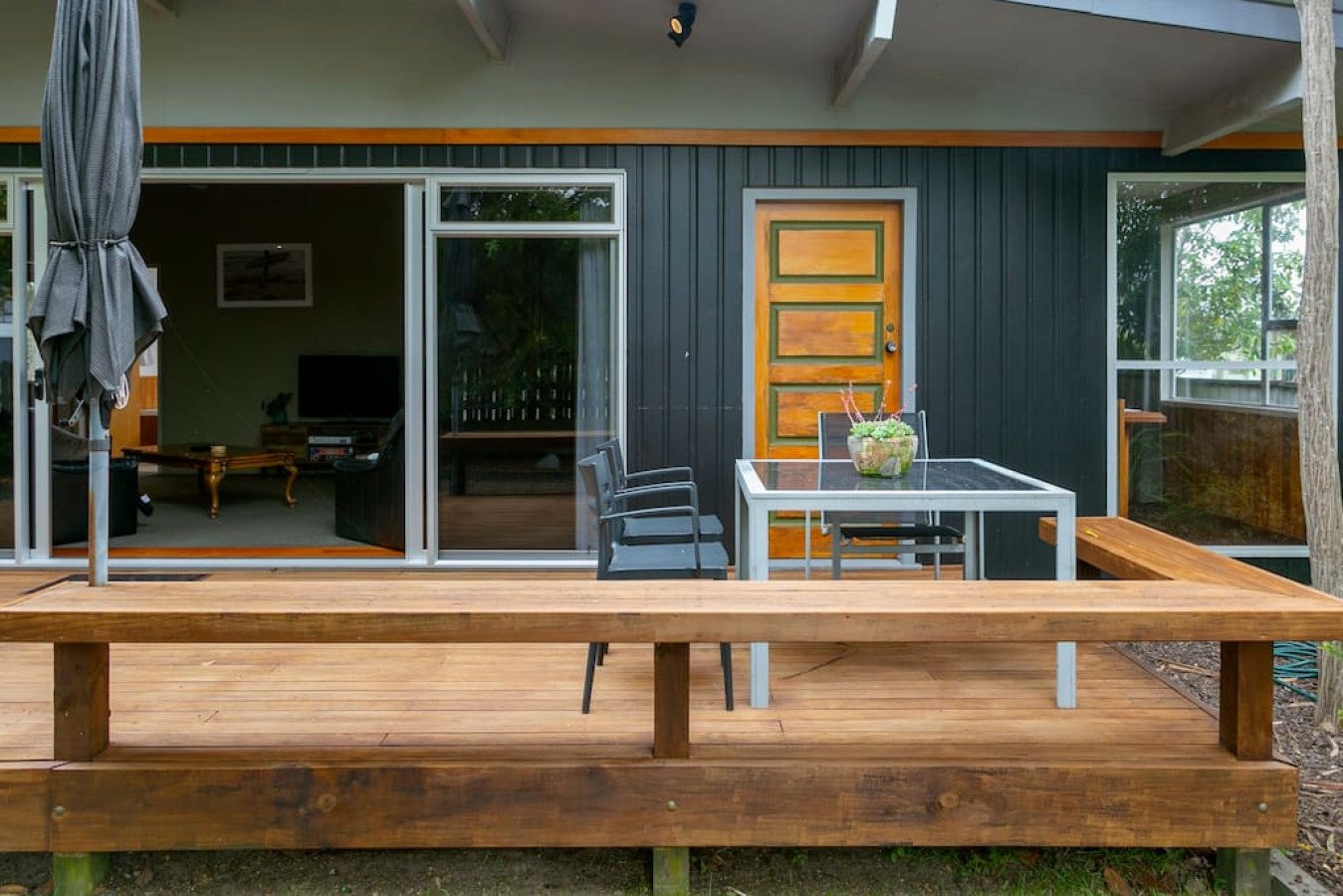 Image of a wooden deck with outdoor seating, including a glass table and three chairs, in front of a house with a black exterior and a yellow wooden door, with sliding glass doors leading to an interior living room.