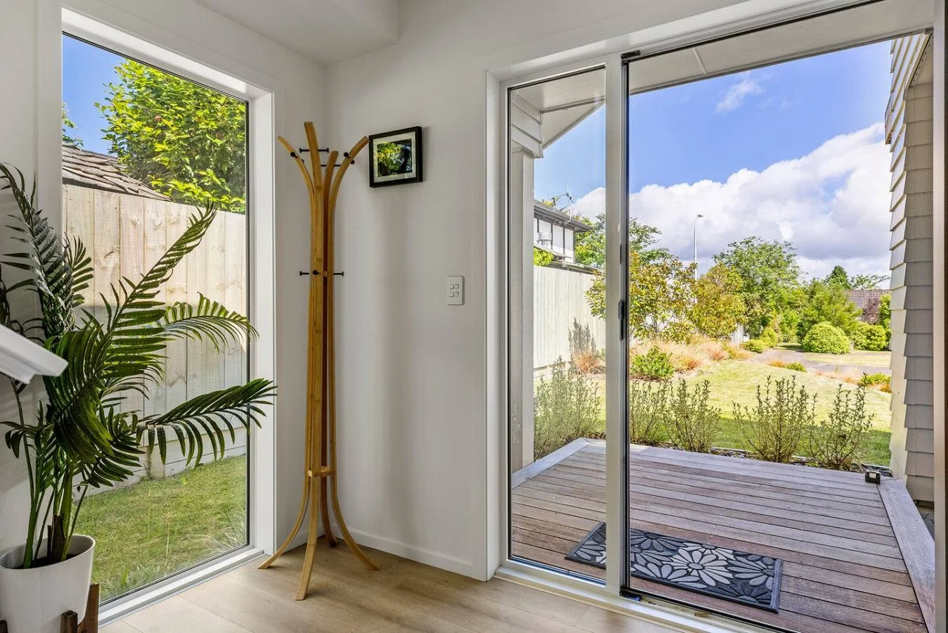 Interior view of a room with a sliding glass door opening to a backyard with a wooden deck, green lawn, shrubs, and trees under a blue sky with clouds.
