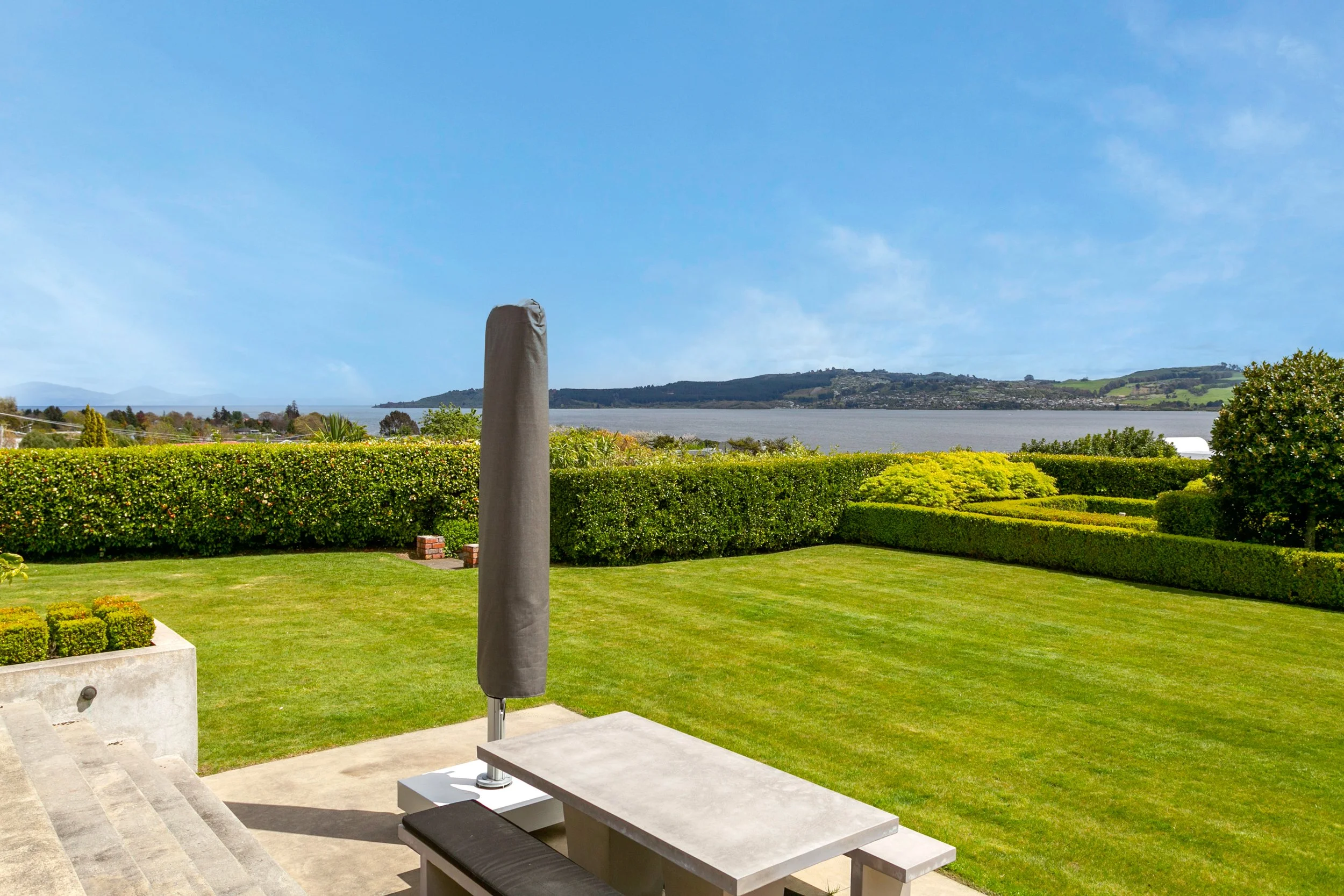 Backyard patio with concrete bench and umbrella, lush green lawn, neatly trimmed hedges, lake and hills in the distance under a clear blue sky.