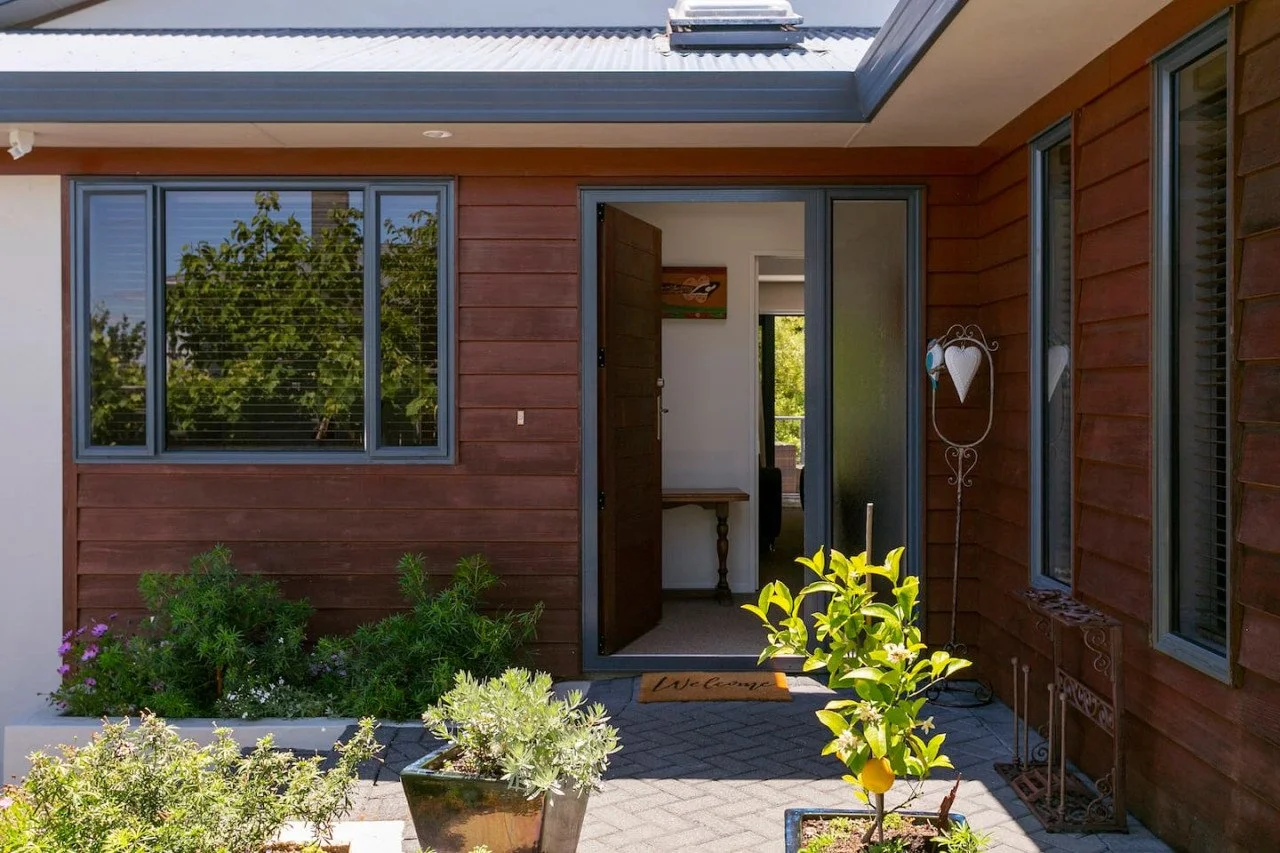 Open front door of a house with wooden exterior, leading into a bright interior. Potted plants and garden decor are visible outside.