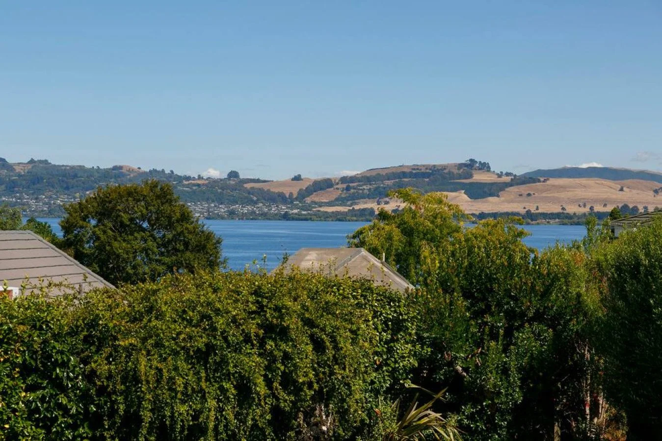 View of a lake with houses and green trees in the foreground, hilly terrain in the background, under a clear blue sky.