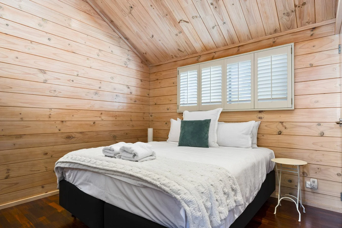 Neatly made bed with white bedding and towels in a small wooden cabin bedroom with wood panel walls and a window with white shutters.