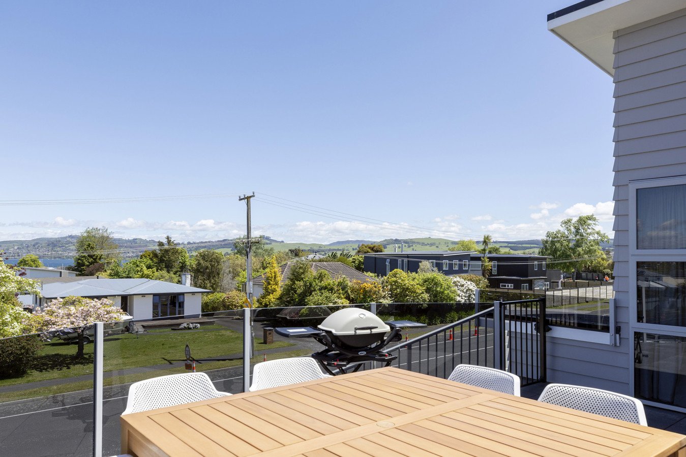 View of a residential neighborhood from a balcony with a wooden dining table, white chairs, and a grill under a clear blue sky.