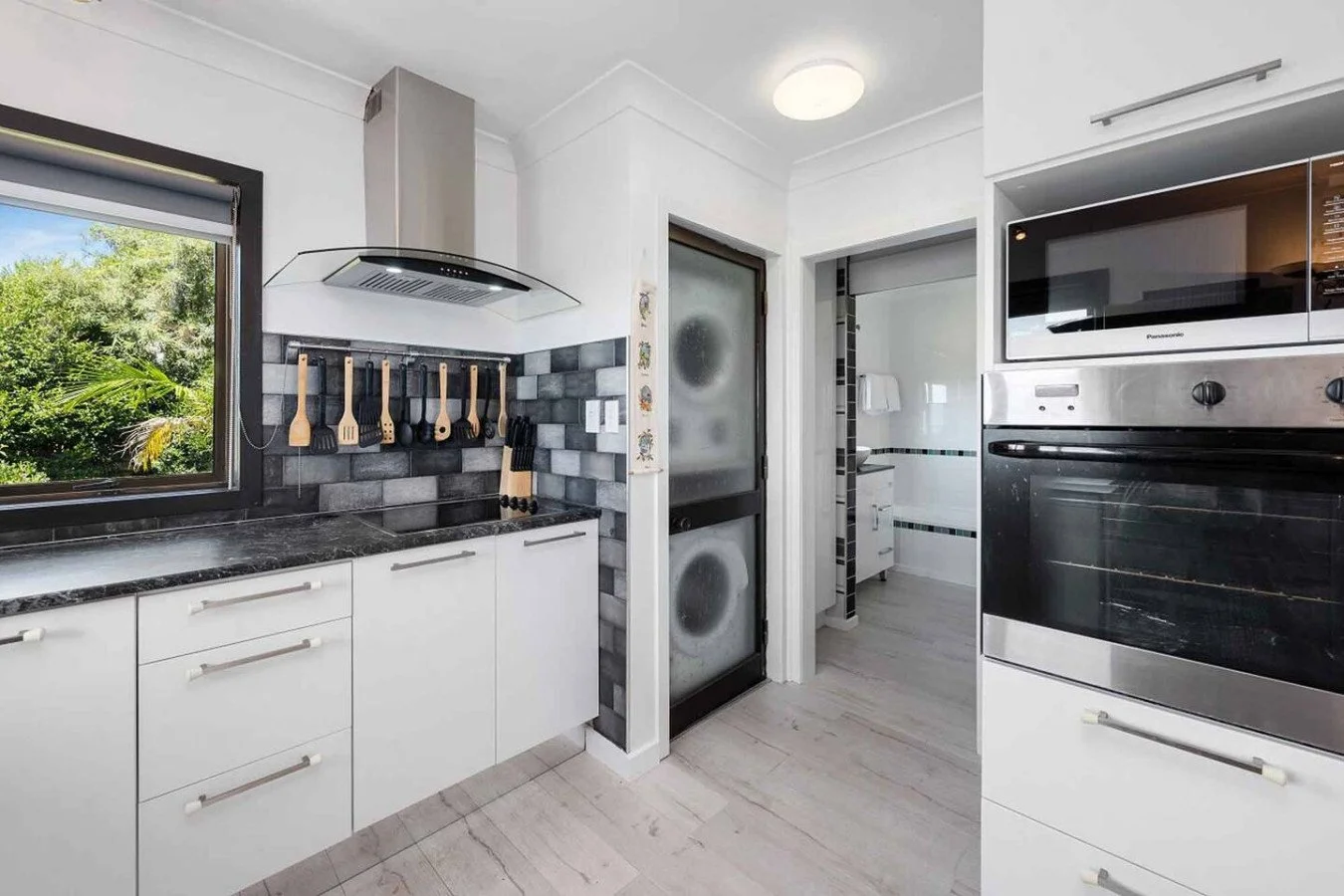 Modern kitchen with white cabinets, black marble countertop, and a large window showing greenery outside. There are wooden utensils hanging above the countertop, a stainless steel range hood, a sliding glass door with a washing machine behind it, and