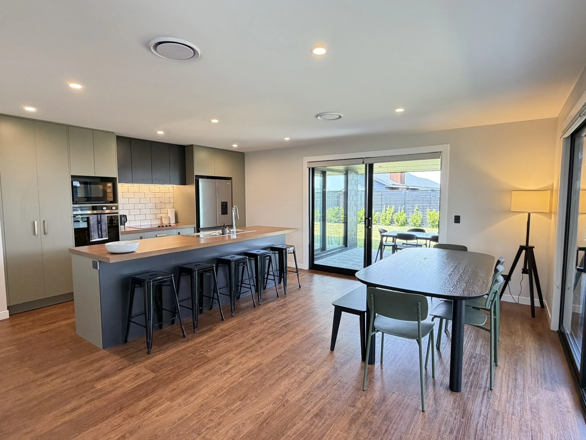 Open-concept kitchen and dining area with hardwood floors, light-colored cabinets, a kitchen island with black stools, large sliding glass doors leading outside, and dining table with chairs near a floor lamp.