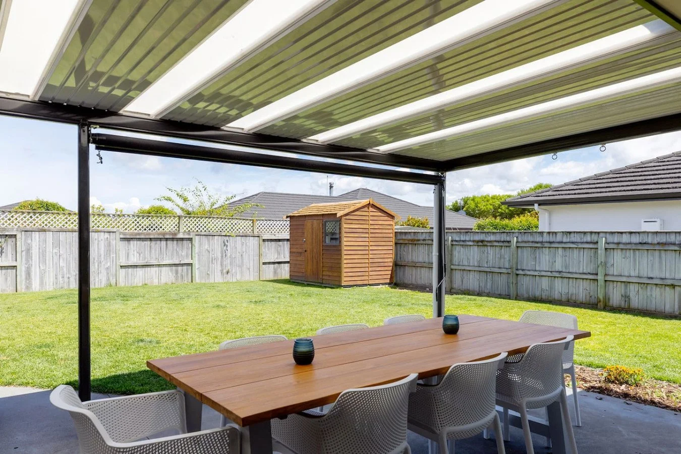 Backyard with a covered patio, wooden dining table, white chairs, green lawn, wooden shed, and wooden fence.