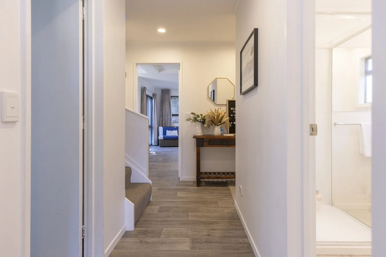 A hallway with wood-look flooring, leading to a living room with large windows, curtains, and a blue sofa. There is a small decorative table with a vase of flowers and wall art.