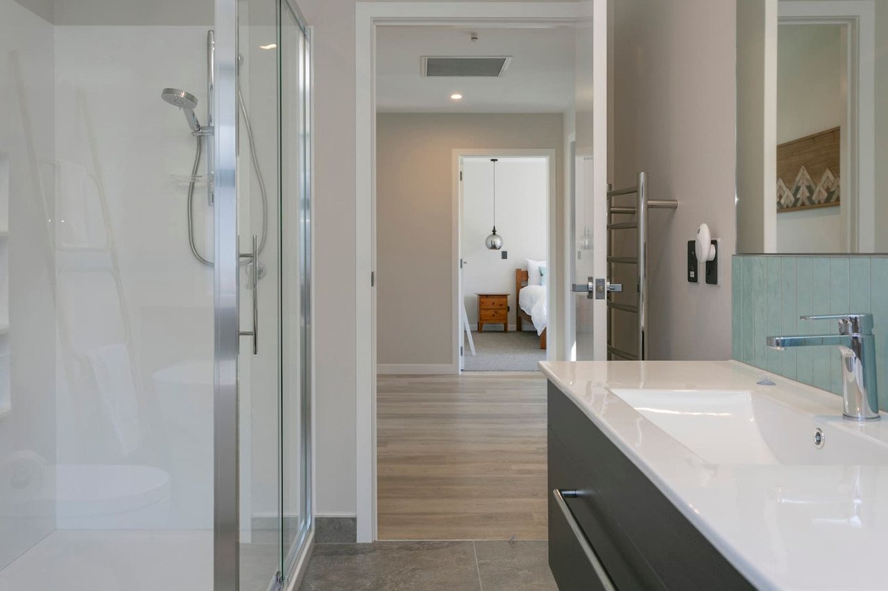 Modern bathroom with a glass shower stall on the left, a sink with a faucet on the right, and a towel warmer on the wall. Through the doorway, a bedroom with a bed, nightstand, and hanging pendant light is visible.