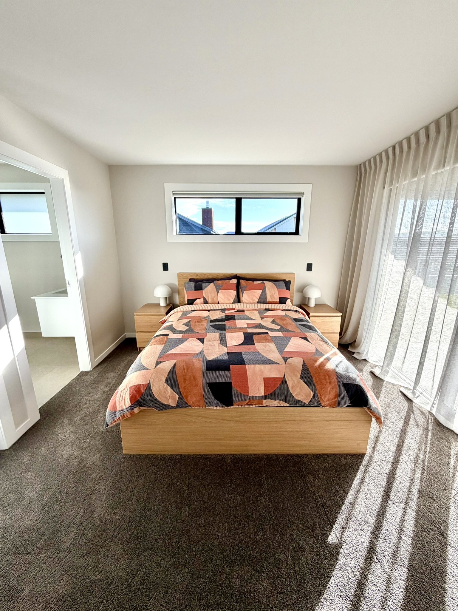 A bedroom with a wooden bed frame, geometric patterned bedspread in shades of pink, orange, black, and beige, two white bedside lamps, a horizontal window, and sheer curtains on the right side. The room has beige carpet and white walls.