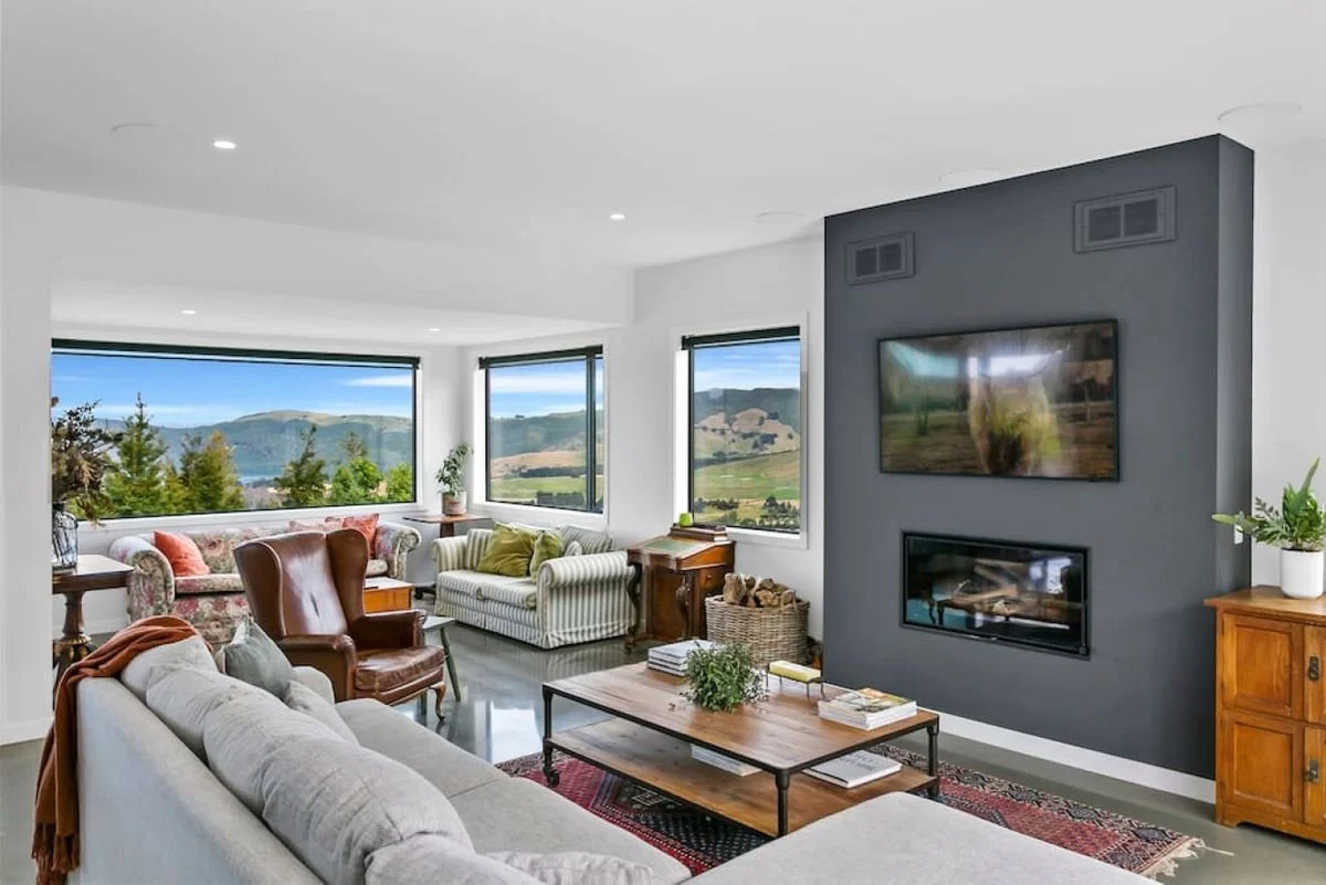 Living room with large windows showing a scenic mountain landscape, featuring a gray fireplace with a TV mounted above, various sofas, armchairs, a wooden coffee table, and potted plants.