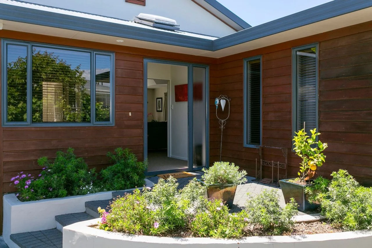 Front entrance of a house with brown wooden siding, gray window frames, and a garden with potted plants and flowering bushes.