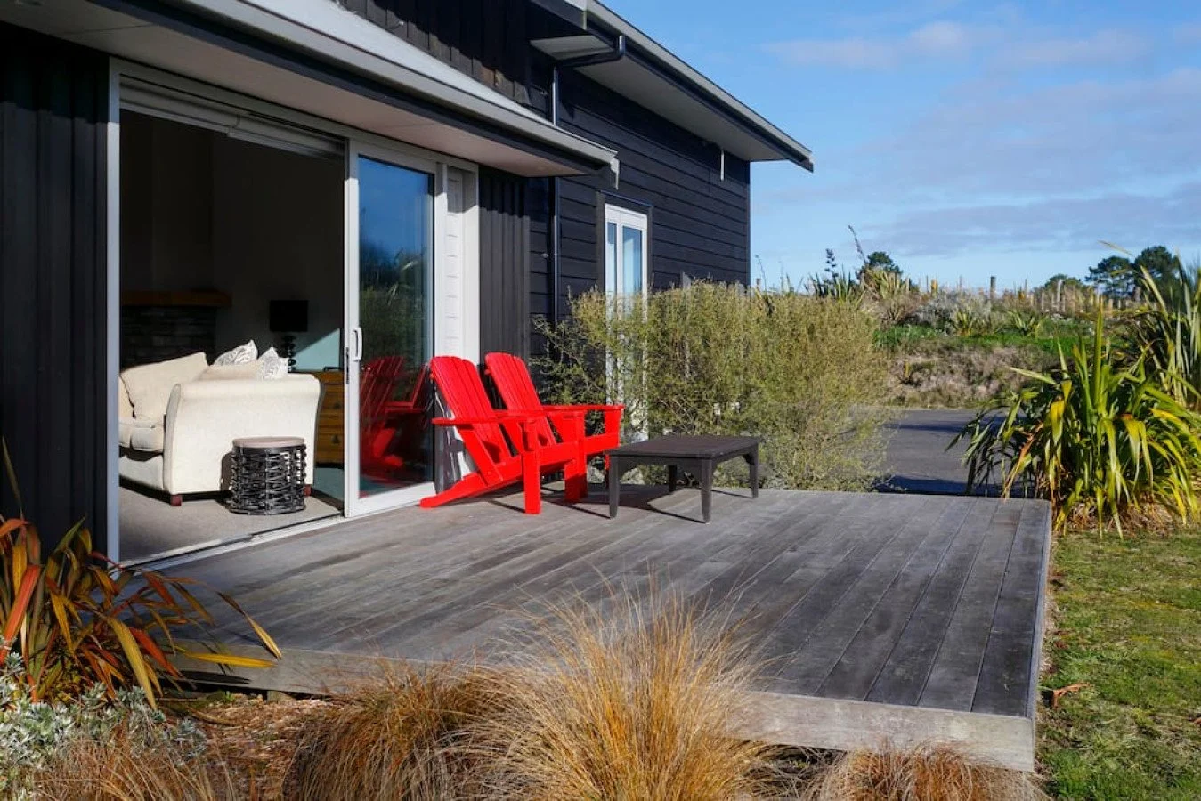 A wooden deck attached to a black house with sliding glass door, outside seating including two red Adirondack chairs, a black side table, and an off-white sofa inside visible through the glass door. Outside plants and bushes surround the deck, with a