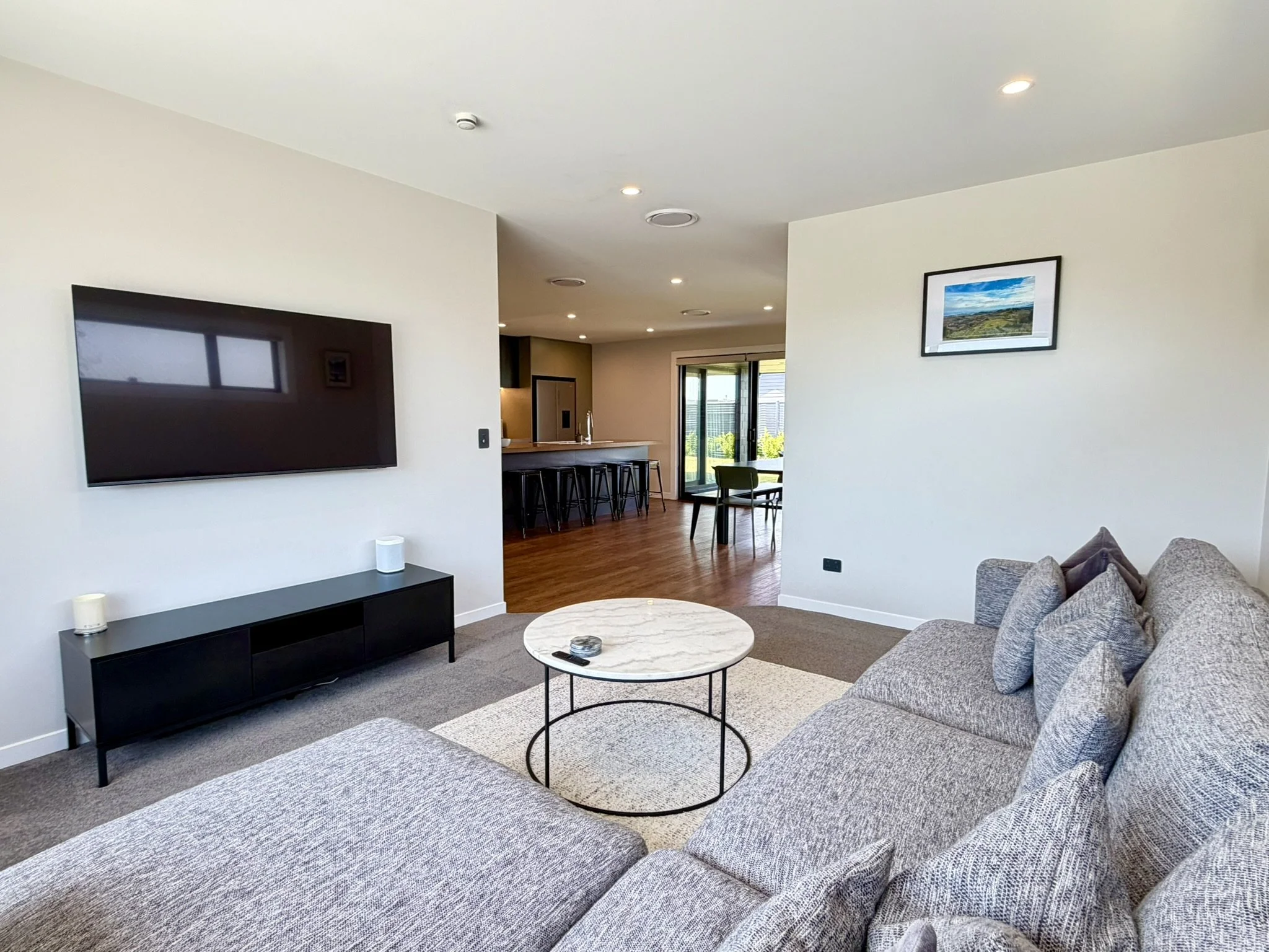 Living room with a wall-mounted TV, a grey sectional sofa, a round marble-top coffee table, a black TV stand, and a wall picture. The room is open to a kitchen and dining area with a sliding glass door leading outside.