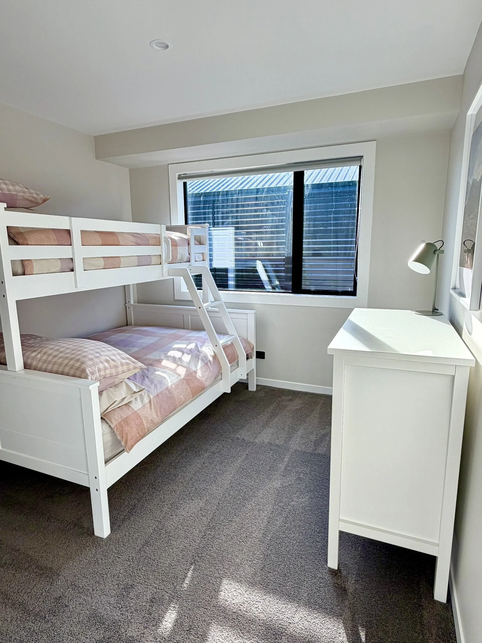 A cozy bedroom featuring white bunk beds with pastel checkered bedding, a window with blinds, a white dresser with a lamp, and a framed picture on the wall.
