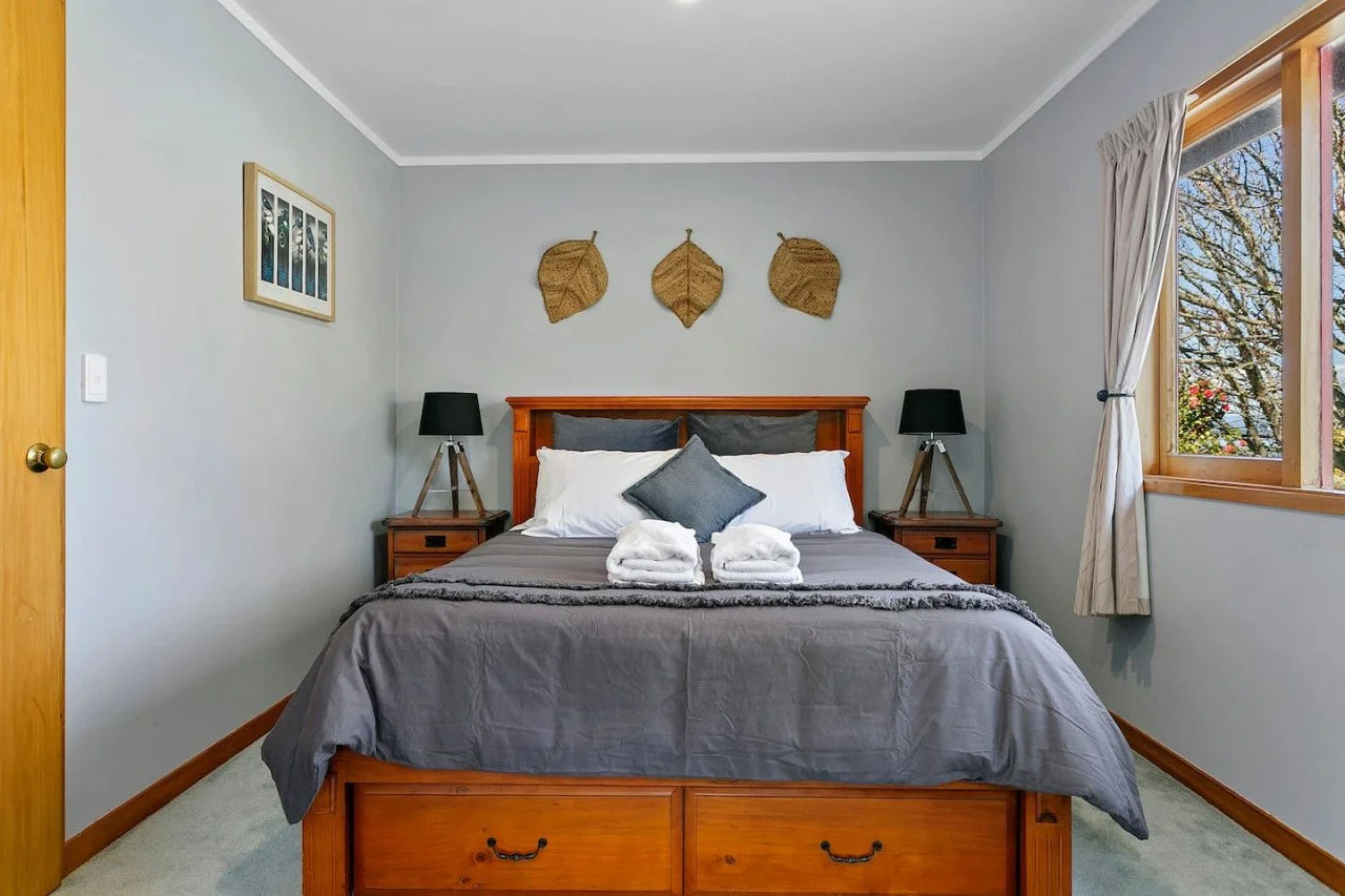 A neatly made bed with gray bedding, white pillows, and two rolled towels, in a bedroom with wooden bedside tables, black lamps, and wall art behind the bed. A window with white curtains shows trees outside.