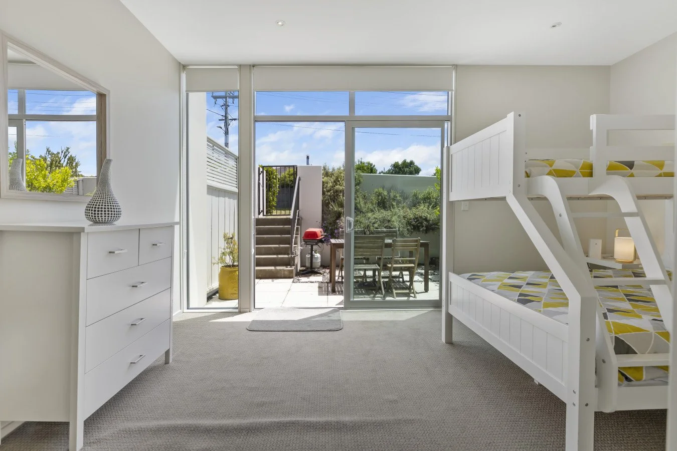 Bright room with white bunk bed, white dresser with decorative vase, and sliding glass door leading to outdoor patio with table, chairs, and a small red grill.