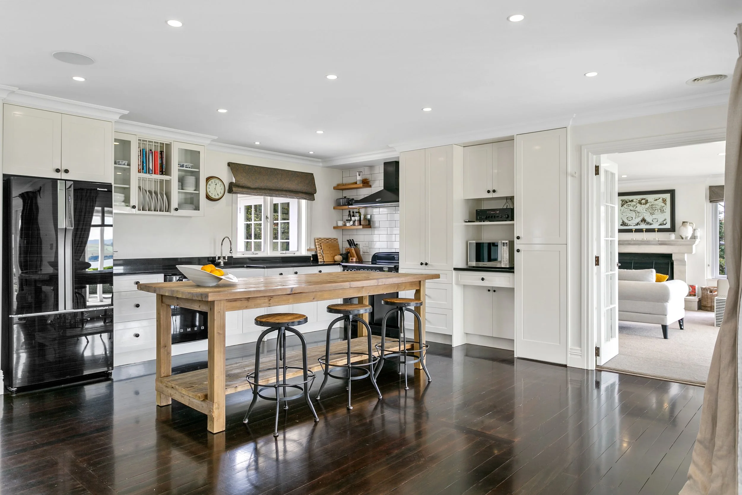 Bright kitchen with white cabinets, black appliances, and a wooden island with three stools, adjacent to a cozy living room with fireplace.