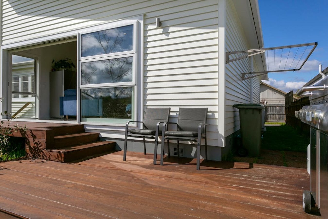 Backyard wooden deck with two chairs, adjacent to a house with sliding glass door, garbage bin, and a barbecue grill, under a blue sky with some clouds.