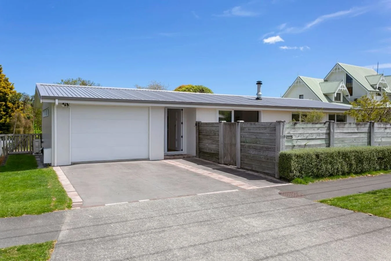 A modern single-story house with a white exterior, grey metal roof, attached garage, concrete driveway, small front yard, wooden fence, and a neighboring multi-story house with pointed rooftops in the background.