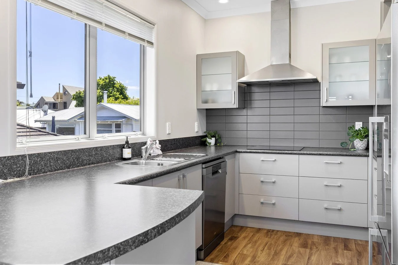 Modern kitchen with gray cabinets, black countertop, stainless steel appliances, and a window overlooking neighboring houses and clear sky.