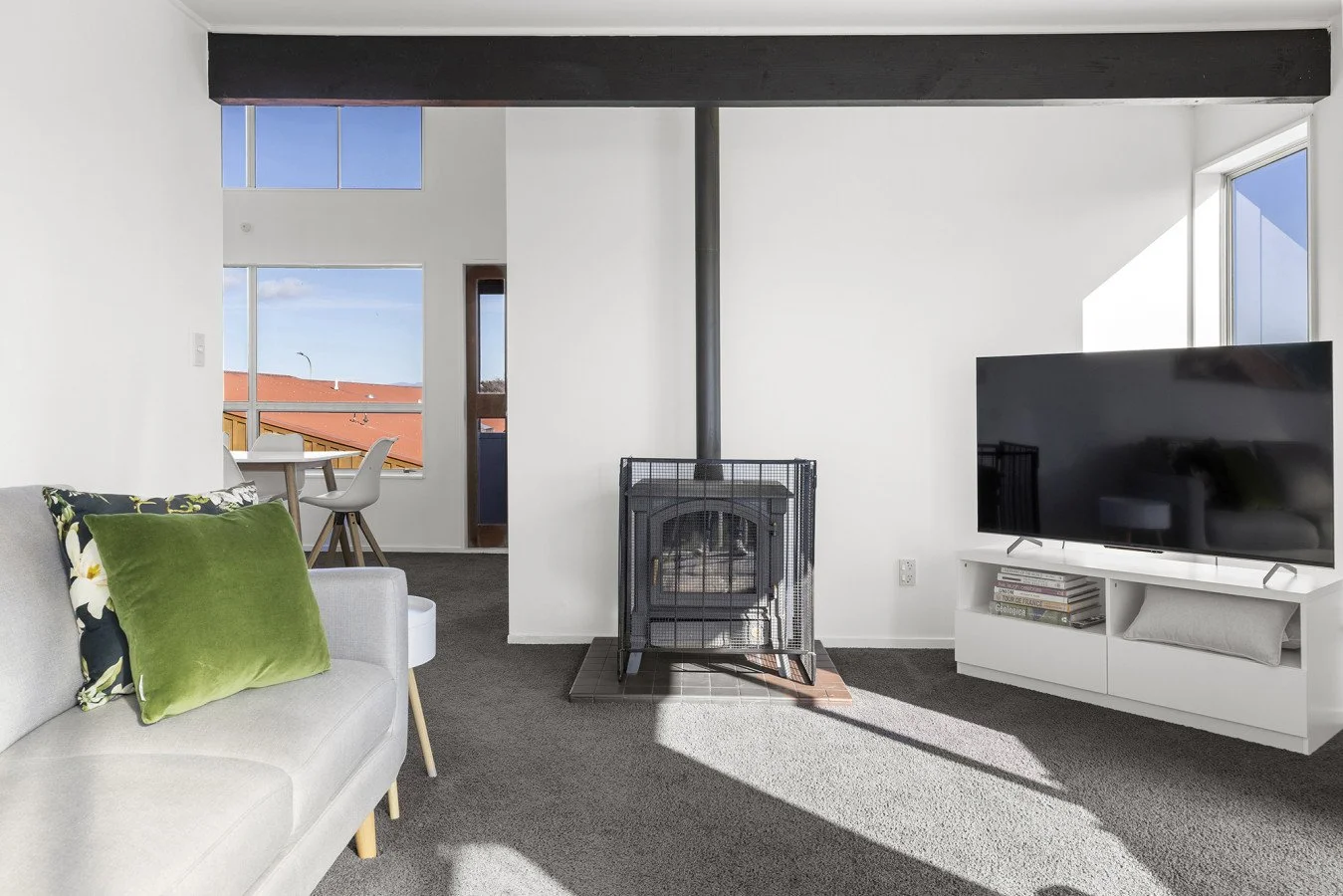 Living room with white walls, gray carpet, a white sofa with green and floral pillows, a white TV stand with a large flat-screen TV, a wood stove with a black protective grate, and sunlight casting shadows on the floor.