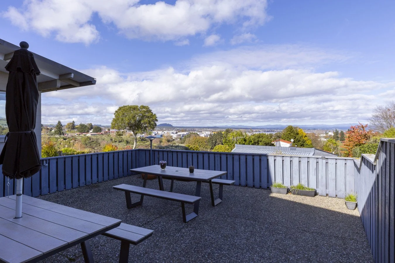 A spacious balcony with a wood picnic table and bench, black potted plants, a closed black umbrella, and a view of trees, rooftops, and mountains under a partly cloudy sky.