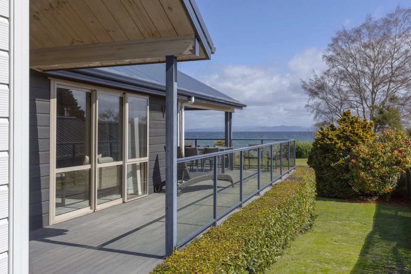 A house with a wooden deck and glass railing, overlooking a body of water with boats, with trees and bushes in the yard.
