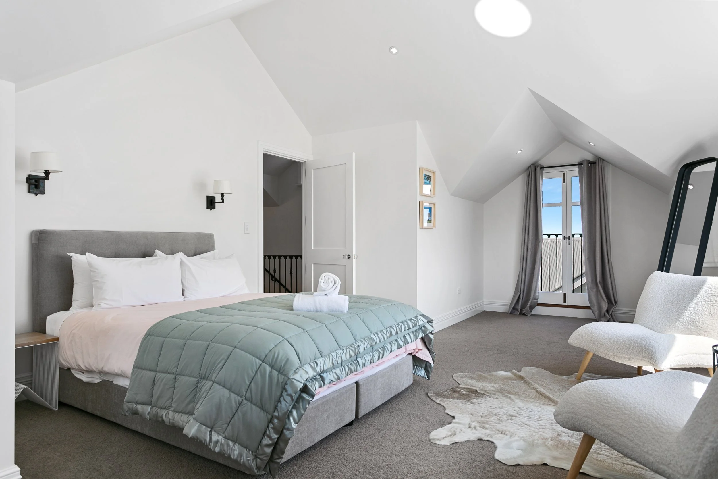 Bright bedroom with a bed, two chairs, a cowhide rug, and a balcony door with curtains.
