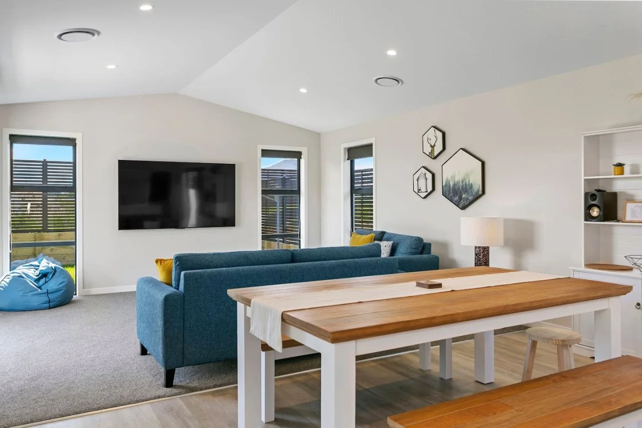 Living room with a mounted flat-screen TV, two windows with blinds, blue sofa, bean bag chair, wall art, bookshelf, and wooden dining table.
